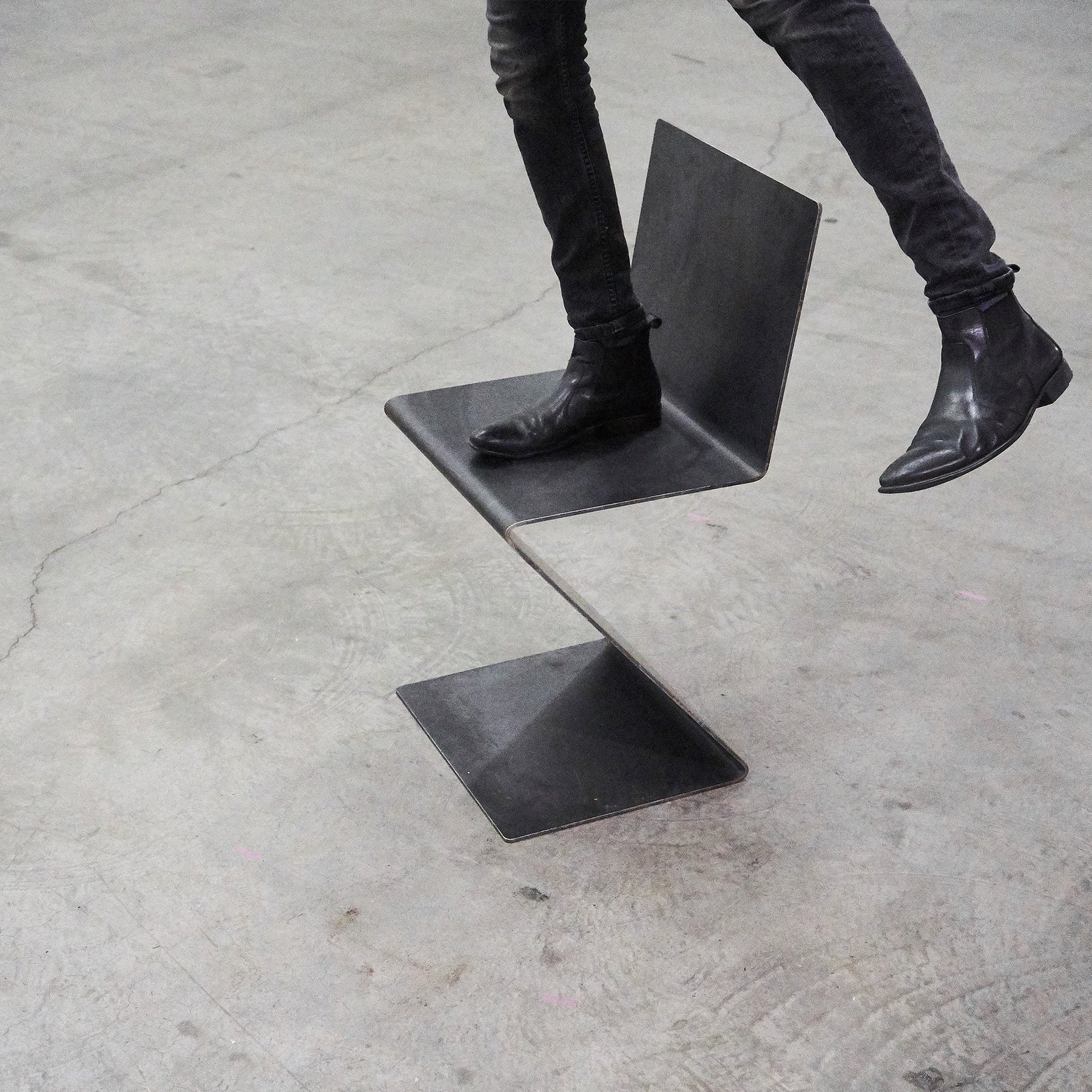 Eine Person steht mit einem Bein auf einem Freischwinger aus rohem Flachstahl auf Betonboden